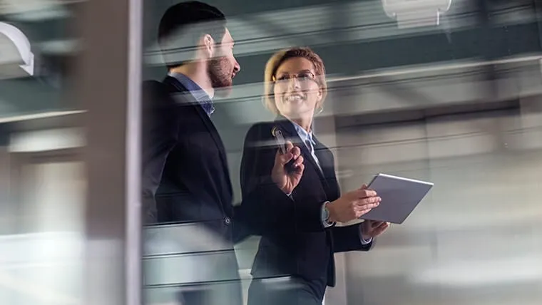 Two business professionals, a woman with a tablet and a man in a suit, discussing while walking in an office corridor.
